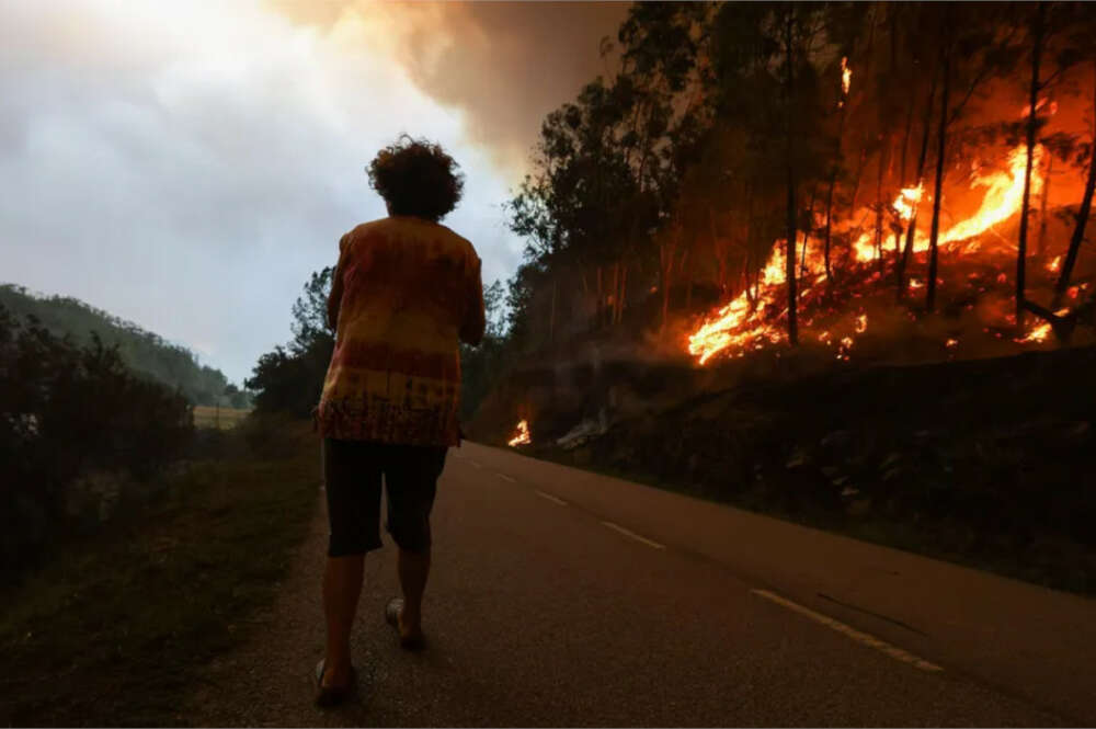 Una mujer camina por la carretera mientras el fuego se extiende por uno de los costados de la vía / Europa Press