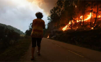 Una mujer camina por la carretera mientras el fuego se extiende por uno de los costados de la vía / Europa Press