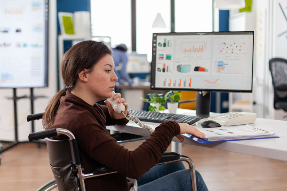 Una mujer en silla de ruedas atendiendo el teléfono mientras tiene una pantalla detrás de computadora